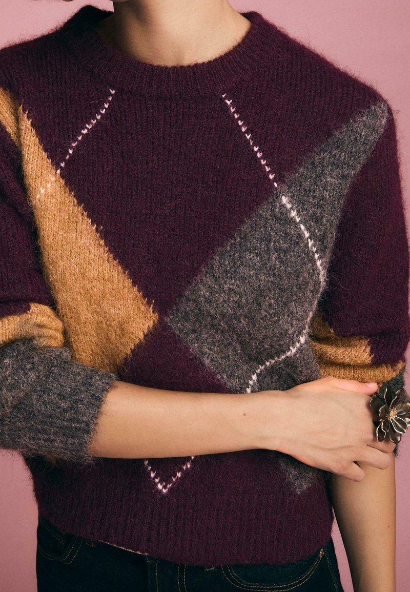 Personne portant un pull en maille duveteuse à blocs de couleurs violet, moutarde et gris, manches retroussées, debout devant un fond rose.