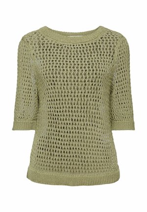 Personne portant un pull beige en maille ajourée à manches courtes par-dessus un haut noir et un pantalon noir taille haute, avec une main derrière le dos.