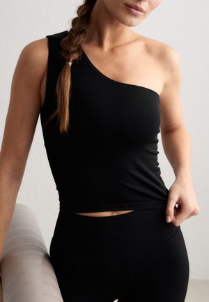 Donna con capelli intrecciati che indossa un top nero monospalla e pantaloni neri, tenendo un tappetino da yoga sotto il braccio contro uno sfondo neutro.