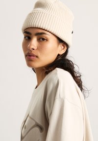 Cream ribbed knit beanie with a slight slouch and subtle logo tag. Worn with a light beige oversized t-shirt, featuring a graphic print.
