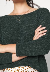 Vihreä neulottu pusero, jossa leveä venepääntie, teksturoitu kangas ja pitkät hihat. Näkyvissä kultaiset kerrokselliset kaulakorut. Kuvioitu hame alapuolella.