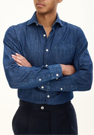 Man with folded arms wearing a blue denim button-up shirt and black pants against a plain background.