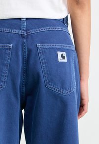 Blauwe spijkerbroek met een gladde textuur, twee achterzakken en een wit logo-embleem. Kenmerkt zichtbare stiksels en een hoge taille.