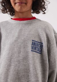 Enfant aux cheveux bouclés portant un sweat-shirt gris avec le logo "AMERICAN VINTAGE" et un sous-pull rouge sur un fond uni.