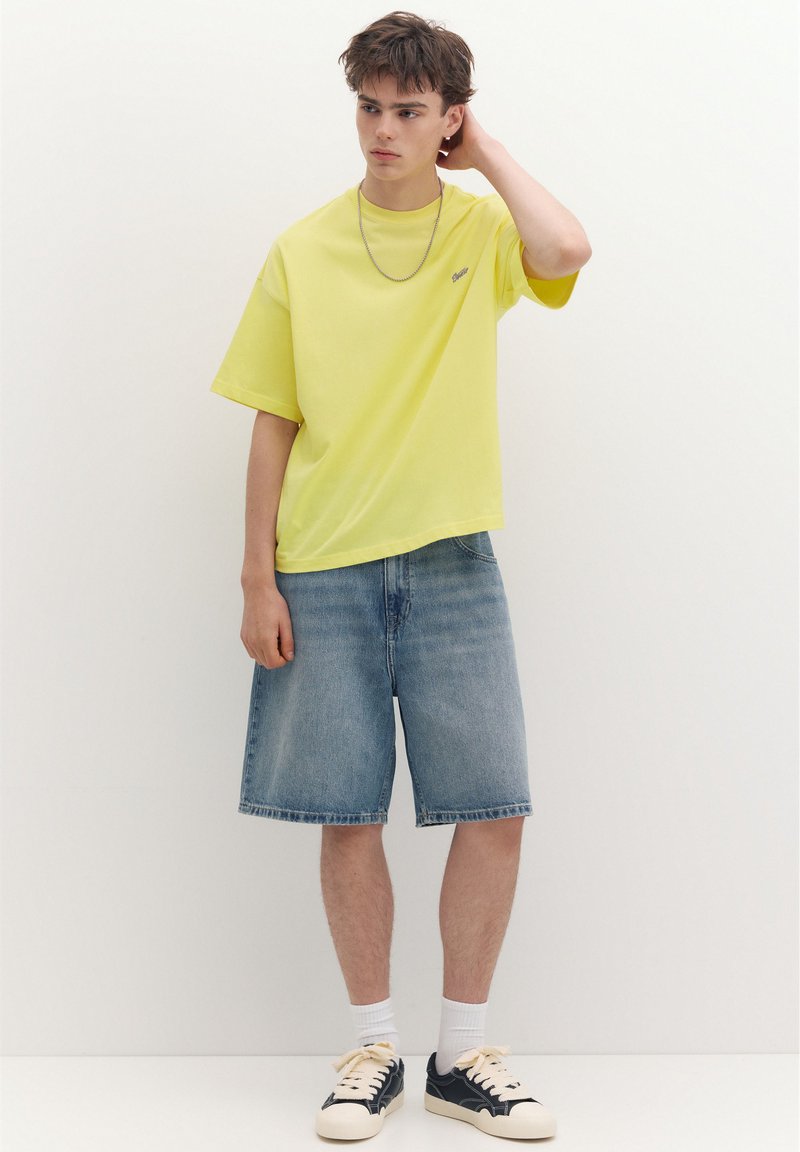 Jeune homme portant un t-shirt jaune, un short en jean jusqu’aux genoux, des chaussettes blanches et des baskets noires et blanches, debout contre un mur blanc uni.