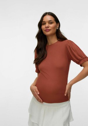 Femme enceinte aux longs cheveux foncés portant un haut côtelé couleur rouille et une jupe blanche, tenant doucement son ventre et souriant.