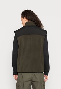 Fleece vest med høy krage i mørkegrønn og svart, med en jevn tekstur og uten ermer. Øvre rygg har en distinkt sømdetalj.
