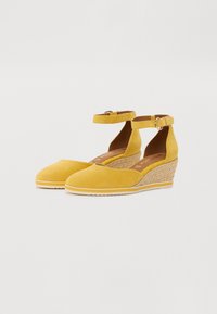 Bottines à talons compensés en suède jaune avec des brides à la cheville, présentant une texture tissée sur le talon compensé, et une semelle lisse. Forme de bout arrondi.