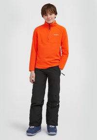 Oranje fleece pullover met een halve rits en hoge kraag, gecombineerd met zwarte sneeuwbroeken en blauwe sneeuwlaarzen met witte veters.