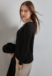 Top nero a maniche lunghe con scollo a barca, realizzato in tessuto morbido. Presenta una vestibilità rilassata e polsini a coste; abbinato a pantaloni beige.