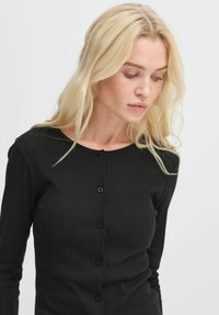 Čierny rebrovaný cardigan s okrúhlym výstrihom a zapínaním na gombíky vpredu. Vyrobený z elastického materiálu, má dlhé rukávy a priliehavý strih.