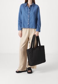 Personne portant une chemise en denim bleu, un pantalon beige à jambes larges, des sandales noires, tenant un grand sac cabas noir matelassé par les poignées.