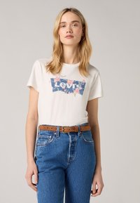 Balta medvilninė marškinė su gėlių Levi's logotipo grafika, derinami su mėlynomis džinsinėmis kelnėmis aukštu liemeniu ir ruduo odiniu diržu.