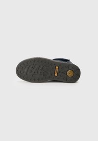 Dark blue fabric shoe with a textured, gray rubber sole featuring star patterns and branding. Accented with a gold emblem.