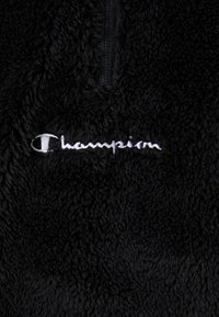 Tissu polaire noir avec une fermeture éclair quart de zip et un logo Champion brodé blanc sur la poitrine.