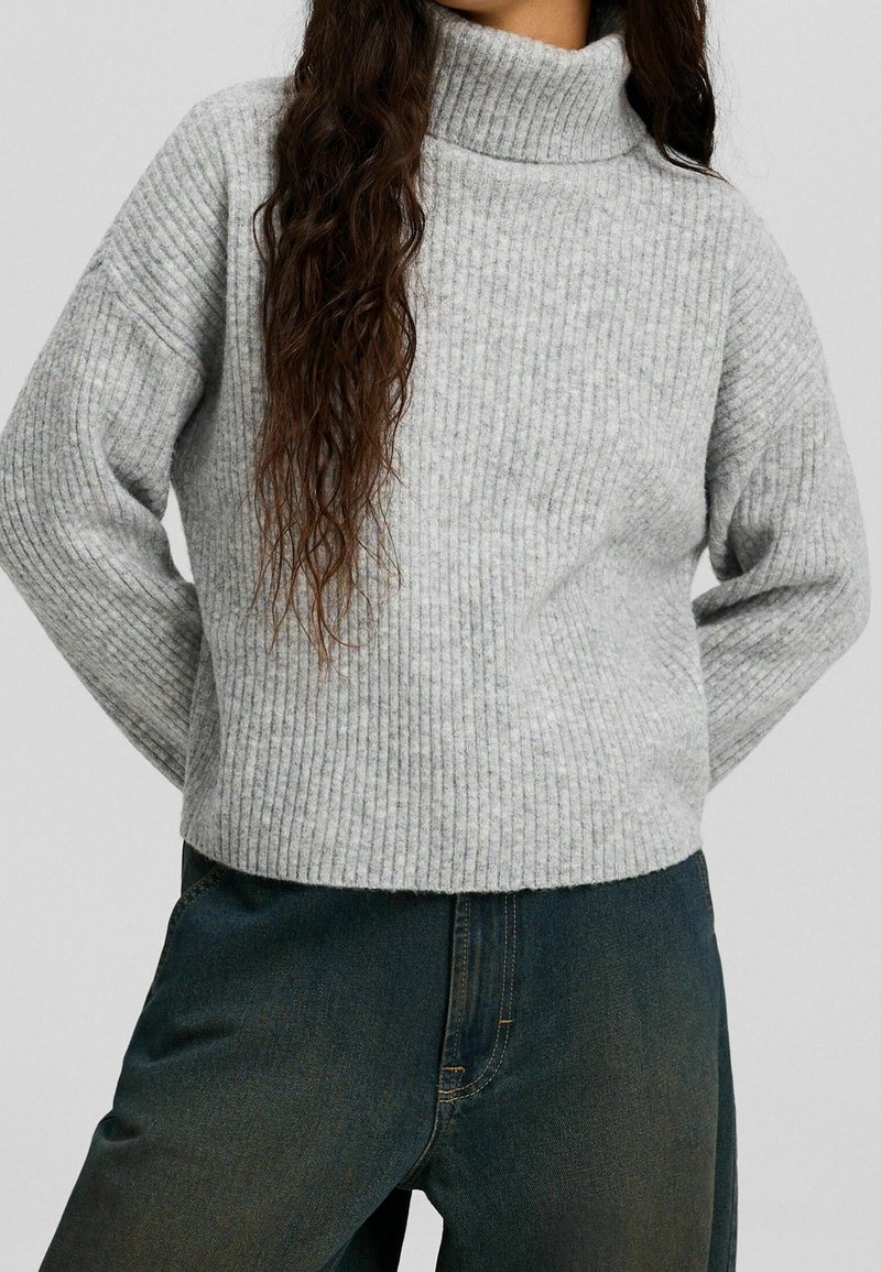 Pull à col roulé gris côtelé à manches longues et coupe décontractée, associé à un jean en denim foncé, mettant en valeur une texture douce.
