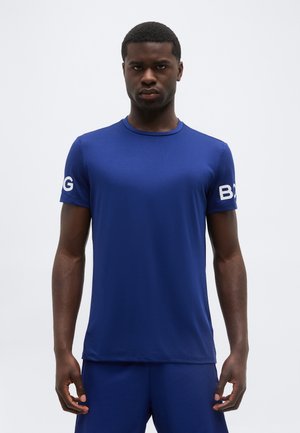 Muž nosiaci priliehavú kráľovskú modrú košeľu s krátkym rukávom s textom „BOSS“ na oboch rukávoch, stojaci pred jednoduchým bielym pozadím.