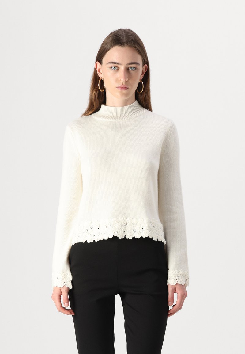 Pull crème avec un col montant, doté d'une bordure en dentelle florale en bas et aux poignets. Fabriqué à partir d'un matériau doux et texturé.