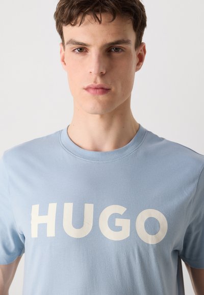 Светло синя памучна тениска с голямо бяло текстово лого "HUGO" отпред. Кръгло деколте и къси ръкави. Гладка текстура.