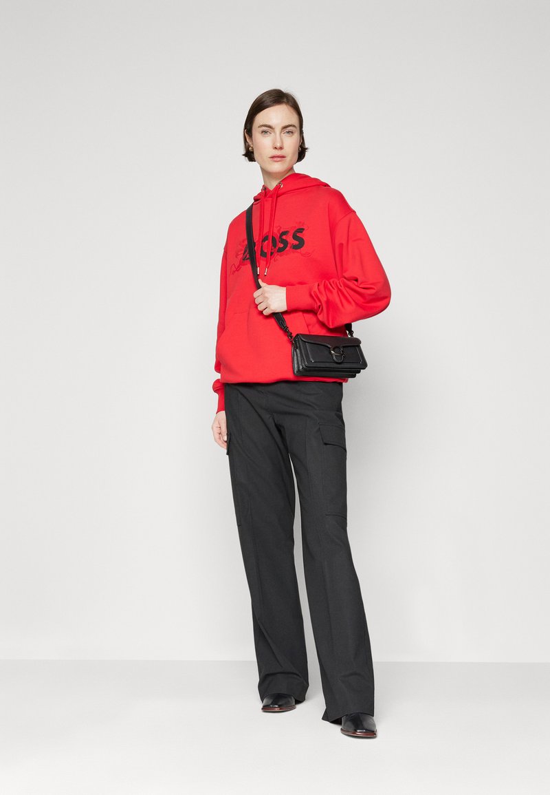 Sweatshirt à capuche rouge avec logo noir, associé à un pantalon large noir. Le modèle tient un petit sac à main noir et porte des ballerines noires.