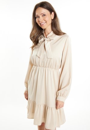 Beige mekko, jossa on solmukkolari, pitkät hihat ja elastinen vyötärö. Siinä on kerroksellinen röyhelöhelma ja sileä pinta.