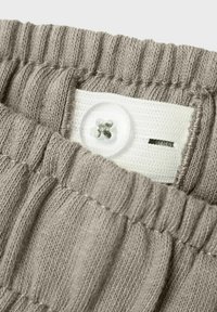 Strikket taupe stof med elastisk tekstur og hvid knapdetalje. Har en smal hvid etiket med en slids til tråd.