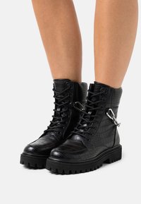 Botines negros hasta el tobillo hechos de cuero sintético con textura de cocodrilo, con cierre de cordones y un acento de hardware plateado en el costado.