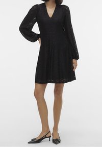 Svart spetsklänning med långa puffärmar, V-ringning och en flödande silhuett. Har ett intrikat blommönster och knälång nederkant.