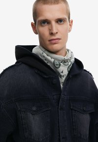 Mörk denimjacka med fransiga axlar, bärs över en svart huvtröja. En lätt scarf med paisleymönster är knuten runt halsen.