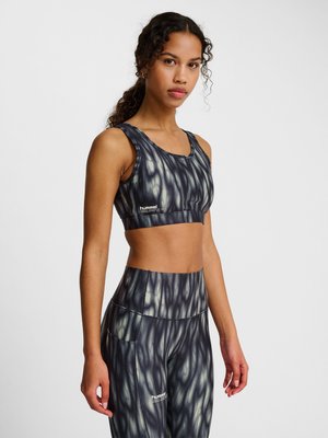 Kvinde iført sort og grå mønstret sports-bh og højtaljede leggings med "hummel"-logo, stående foran en ensfarvet lys baggrund.