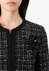 Frau trägt eine schwarz-weiß gemusterte Tweedjacke mit einer schwarzen Stoffblumenbrosche auf der linken Brust.