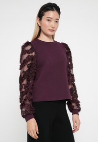 Violeta džemperis ar caurspīdīgām rīvētu piedurknēm. Džempera ķermenis ir vienkrāsains, bet piedurknes ir rotātas ar ziedu motīviem. Rīvēti elastīgie gali noslēdz dizainu.