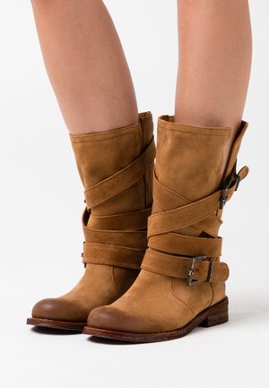 Bottes cheville en daim marron avec des sangles croisées à boucles métalliques, un bout arrondi et un petit talon empilé. Texture lisse avec une finition mate.