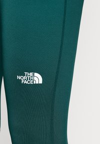 Zielone legginsy sportowe z gładkiego materiału, z białym logo "THE NORTH FACE" oraz wyraźnym detalem szwu.