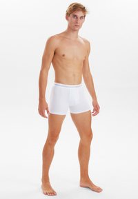 Boxer da uomo bianchi con tessuto liscio, design aderente e elastico visibile in cima. Il modello è in piedi a piedi nudi contro uno sfondo neutro.