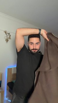 Uomo con barba e maglietta nera che solleva una giacca marrone all'interno, camera da letto con letto e luce LED blu sullo sfondo.