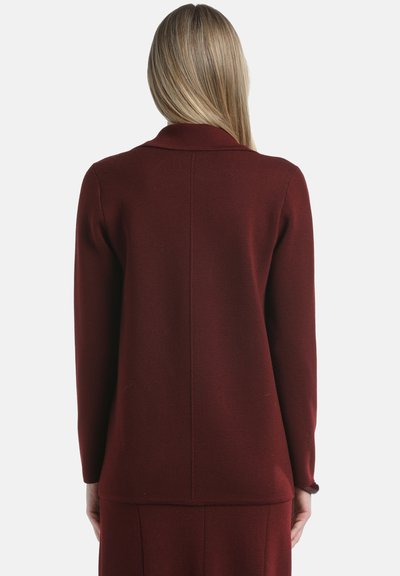 Woman with long blonde hair wearing a dark red long-sleeve jacket and matching skirt, standing facing away against a plain background.