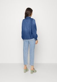 Blå denimskjorta med krage och långa ärmar, i kombination med ljusblå jeans och gröna klumpiga klackar. Huvudfokus ligger på skjortan.