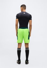 Νεαρός αθλητής φορώντας μαύρο συμπιεστικό μπλουζάκι, αθλητικά σορτς νέον πράσινου χρώματος, μαύρες κάλτσες μέχρι το γόνατο και παπούτσια ποδοσφαίρου, στέκεται γυρισμένος με την πλάτη σε λευκό φόντο.