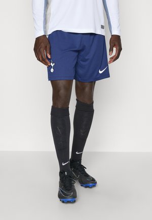 Mørkeblå fodboldshorts med et Spurs-emblem, parret med sorte Nike-sokker og støvler med blå detaljer. Fodboldtrøjen er hvid.