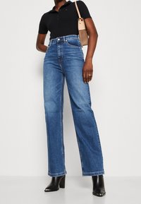Jeans de mezclilla de cintura alta y pierna ancha de color azul, con dobladillo sin terminar, combinados con una camiseta polo negra y botas negras de tacón.