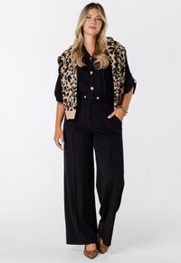 Vrouw in zwarte wijde-pantalon en top met gouden knopen, met een luipaardprint trui over de schouders en bijpassende luipaardprint hakken.