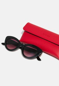 Des lunettes de soleil noires en forme d'œil de chat avec des verres dégradés roses, posées à côté d'un étui texturé rouge, marqué "GUESS" avec une fermeture rabattable.