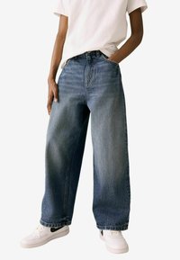 Tumedas sinine teksapüksid kõrge vöökohaga ja laia säärega, millel on hele pleekimine, klassikalise viie tasku disain ning nööpidega sulgemine.