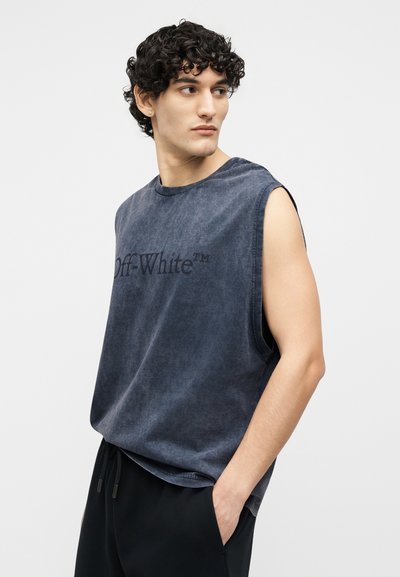 Νεαρός με σγουρά σκουρόχρωμα μαλλιά που φοράει ένα μαν sleeveless γκρι ανθρακί πουκάμισο Off-White και μαύρο παντελόνι, κοιτάζει προς τα αριστερά του.