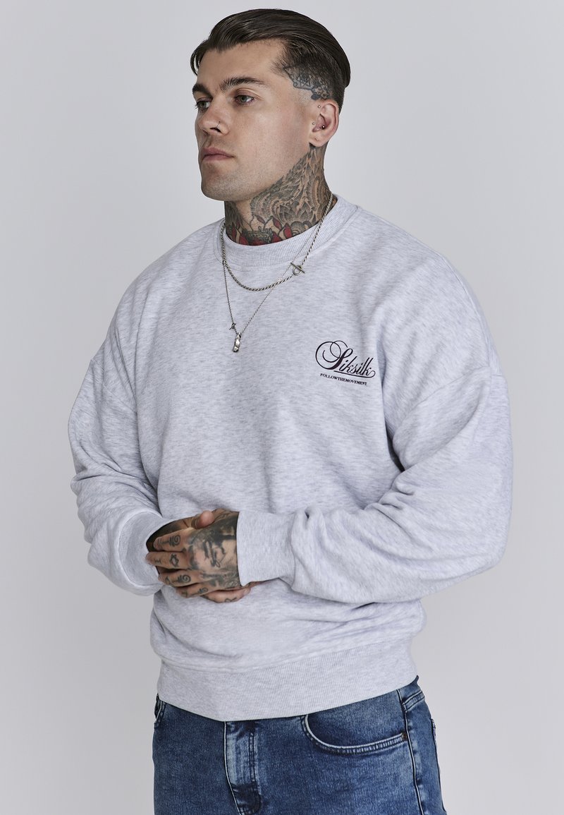 Hellgrauer Pullover mit gerippten Bündchen und Saum, der ein kleines dunkles Logo auf der Brust trägt. Das Modell hat Tattoos und trägt mehrere Lagen von Halsketten.