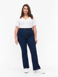 Jeans altos de cintura en azul con forma acampanada, combinados con una camiseta de cuello en V blanca. Zapatillas blancas completan el conjunto. Fondo liso.