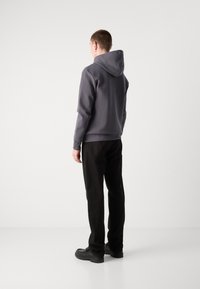 Sudadera gris con capucha, de mangas largas y puños acanalados, combinada con pantalones negros y zapatos negros, sobre un fondo neutro.