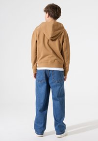 Sudadera con capucha marrón, material de algodón, corte relajado. Combinada con unos vaqueros azules de corte holgado. Diseño simple, sin patrones ni elementos metálicos visibles.