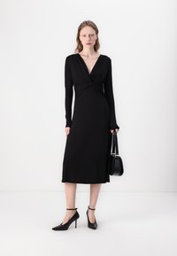 Black long-sleeve dress with a deep V-neck and twisted front detail, knee-length, made from a smooth fabric. Worn with black heels and handbag.